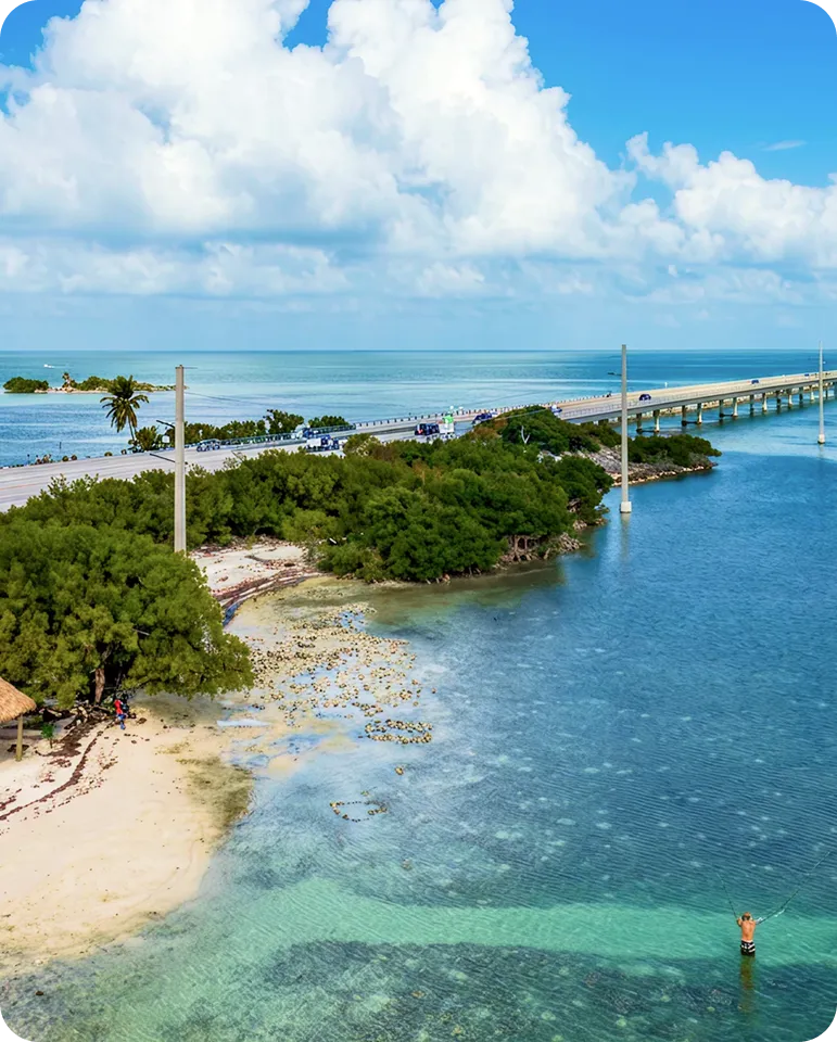 Aerial view of a coastal bridge over turquoise water with sandy shore and green vegetation.