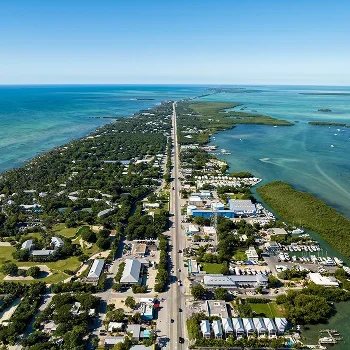 Aerial view of a coastal road flanked by buildings, greenery, and blue ocean waters under a clear sky.