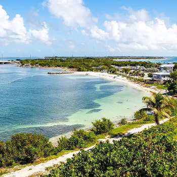 A coastal beach scene with clear blue water, white sand, green vegetation, and scattered buildings under a partly cloudy sky.