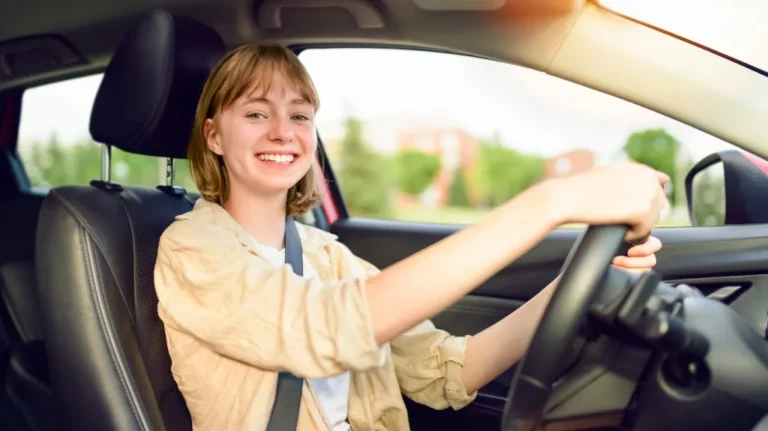 Young woman with short blonde hair smiling while driving a car, wearing a beige jacket and seatbelt.