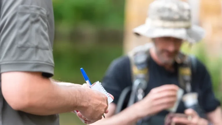 Close-up of a person writing with a blue pen on a small notepad, another person blurred in background.