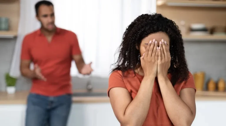 Woman in orange shirt covering face with hands, man in background gesturing.