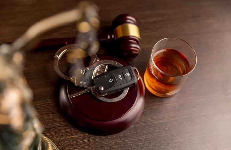 Close-up of a wooden judge's gavel with handcuffs, car keys, and a glass of amber liquid on a wooden surface.