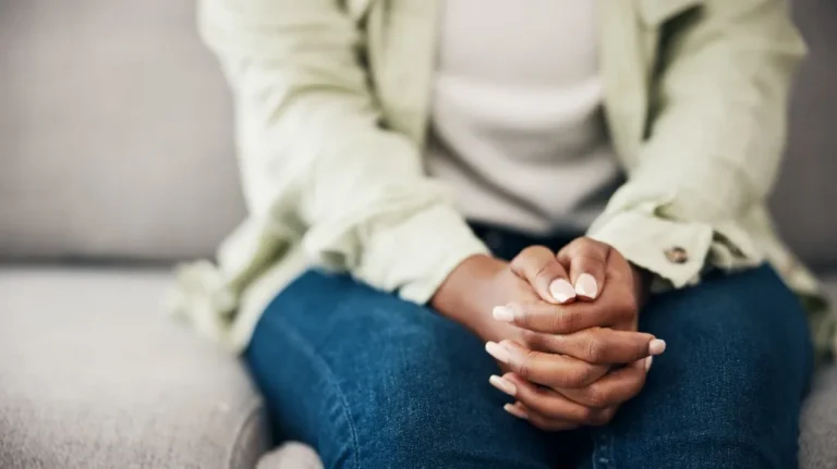 Close-up of a person with interlaced fingers resting on their lap, wearing blue jeans and a light-colored jacket.