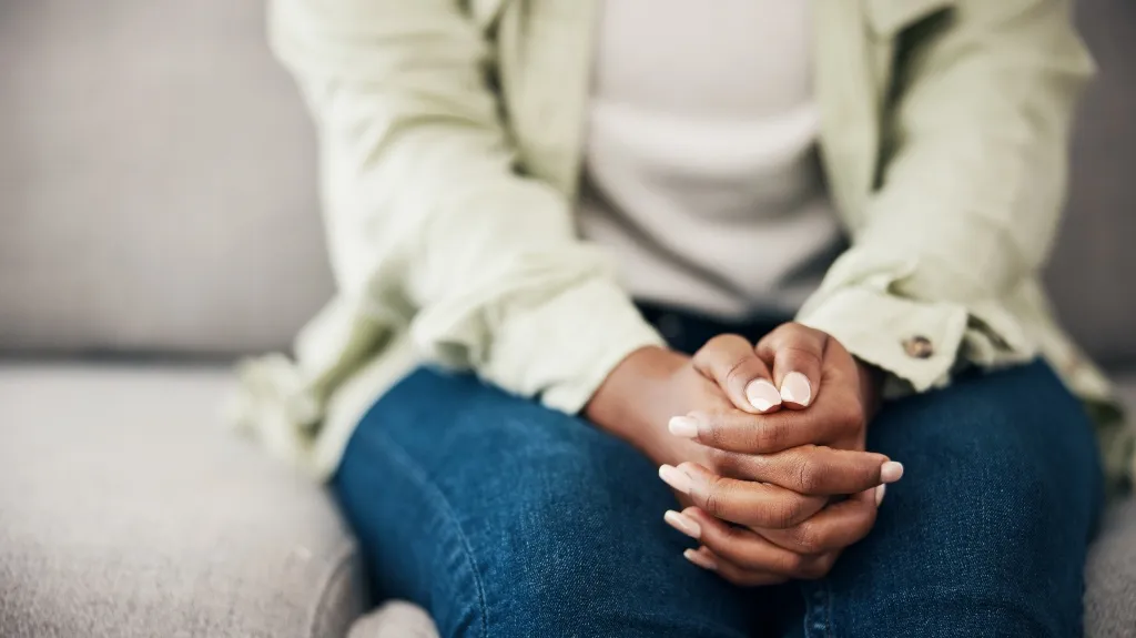 Close-up of a person with interlaced fingers resting on their lap, wearing blue jeans and a light-colored jacket.