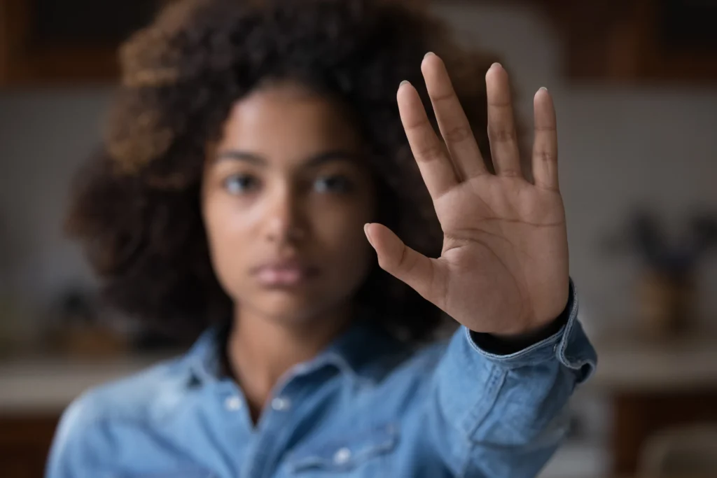 Young woman in a blue shirt with curly hair holding up her hand towards the camera.