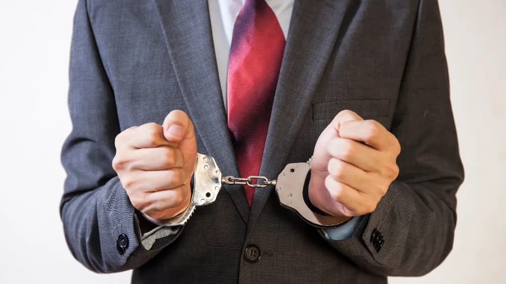 Man in gray suit and red tie with hands cuffed in silver handcuffs.