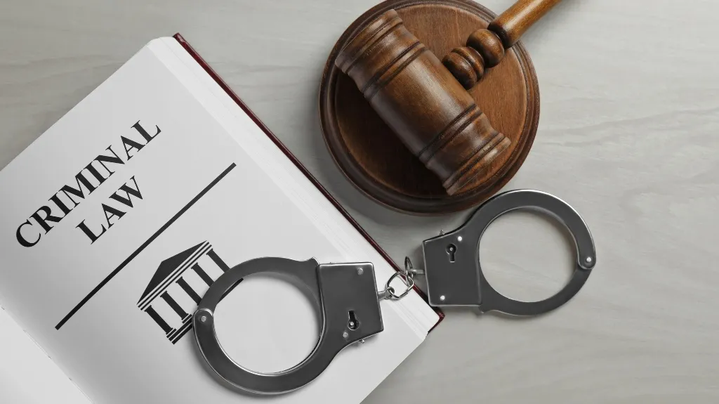 Open book titled "Criminal Law" with metal handcuffs and wooden judge's gavel on a gray surface.